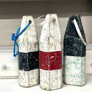 May include: Three white, black, and red nautical buoy floats with rope handles. The buoys are weathered and have a distressed look.