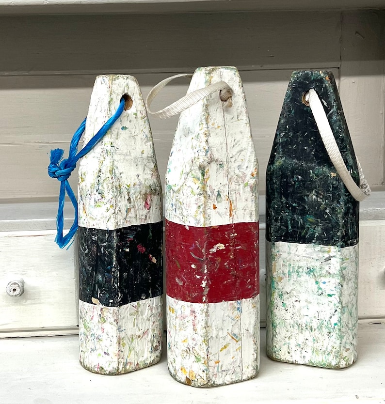 May include: Three white, black, and red nautical buoy floats with rope handles. The buoys are weathered and have a distressed look.