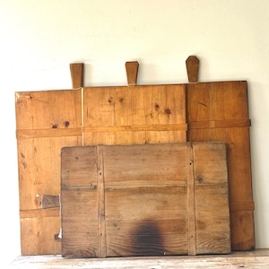 May include: Three antique wooden cutting boards of varying sizes. The boards are made of light brown wood with visible grain and knots. Each board has a handle. The boards are stacked against a white wall.