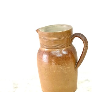 May include: A brown ceramic pitcher with a wide mouth and a handle. The pitcher has a glazed finish and is decorated with horizontal lines.