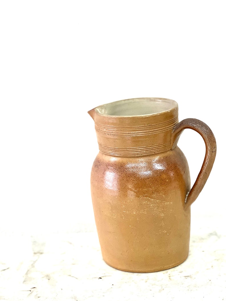 May include: A brown ceramic pitcher with a wide mouth and a handle. The pitcher has a glazed finish and is decorated with horizontal lines.
