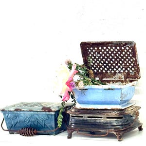 May include: Vintage blue metal containers with a distressed finish. One container has a hinged, perforated lid and is filled with flowers. The containers are stacked on a decorative metal base, showcasing a rustic aesthetic.