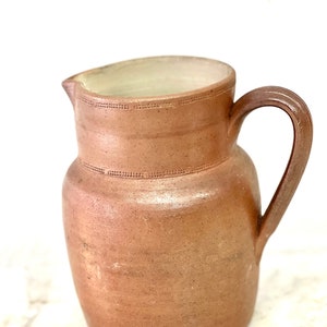 May include: A brown earthenware pitcher with a wide mouth and a handle. The pitcher has a textured surface and a slightly off-white rim.