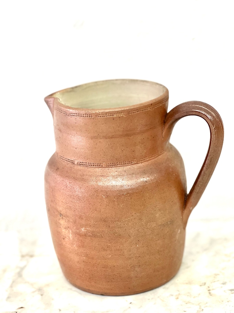 May include: A brown earthenware pitcher with a wide mouth and a handle. The pitcher has a textured surface and a slightly off-white rim.