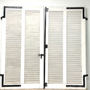 Puede incluir: Un par de contraventanas de madera blanca con lamas horizontales. Bisagras y escuadras de esquina de metal negro aseguran las contraventanas. Un pestillo negro está centrado en la contraventana derecha. Fondo blanco.