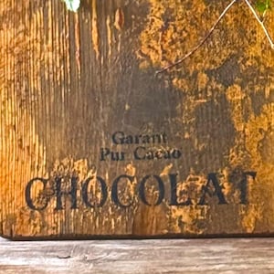 Puede incluir: Letrero de madera con la palabra "CHOCOLAT" en letras negras grandes. El letrero también tiene las palabras "Garant Pur Cacao" en letras más pequeñas. La madera tiene un aspecto envejecido y desgastado con vetas y variaciones de color visibles.