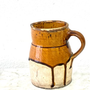 May include: A brown and white ceramic pitcher with a handle. The pitcher has a brown glaze with drips of glaze running down the side.