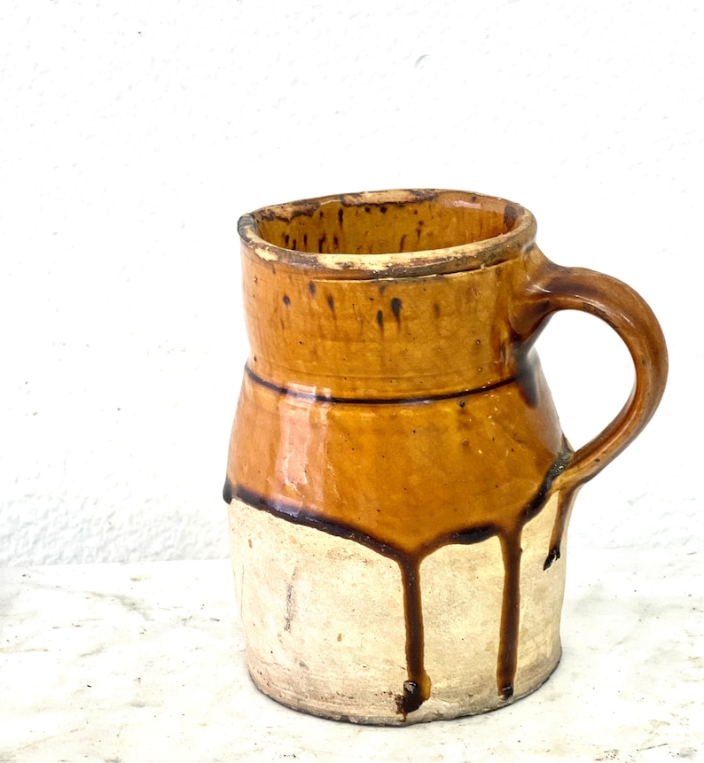 May include: A brown and white ceramic pitcher with a handle. The pitcher has a brown glaze with drips of glaze running down the side.
