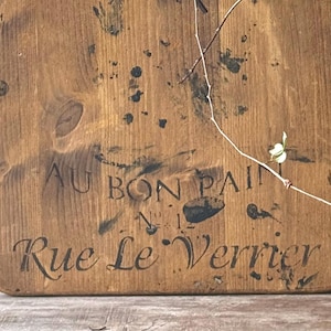 Puede incluir: Una tabla de cortar rectangular de madera con el texto "AU BON PAIN N°1 Rue Le Verrier" en letras negras. La tabla tiene un aspecto rústico y desgastado con manchas oscuras y una veta de madera natural. Una ramita con pequeñas hojas verdes está sobre la tabla.