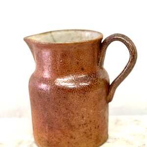 May include: A brown ceramic pitcher with a white interior and a handle. The pitcher has a textured surface and a slightly rounded shape.