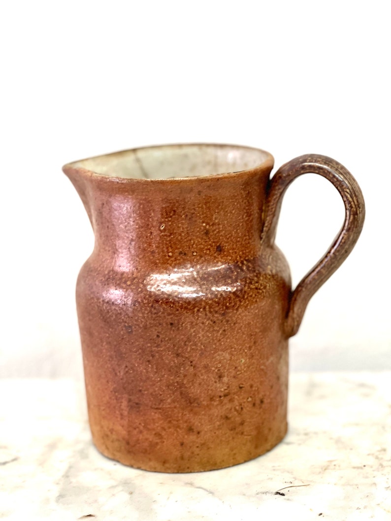 May include: A brown ceramic pitcher with a white interior and a handle. The pitcher has a textured surface and a slightly rounded shape.
