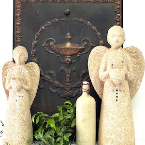 May include: Two beige angel statues with outstretched wings flank a vintage bottle and greenery. A dark, ornate metal panel with a decorative design serves as a backdrop. The angels hold small objects, and the bottle has a wire closure.