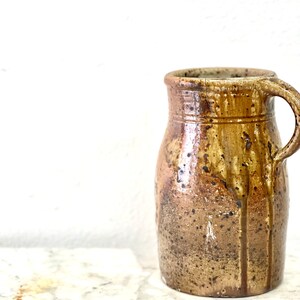 May include: A brown ceramic pitcher with a speckled glaze and a handle. The pitcher has a rounded body and a slightly flared lip.