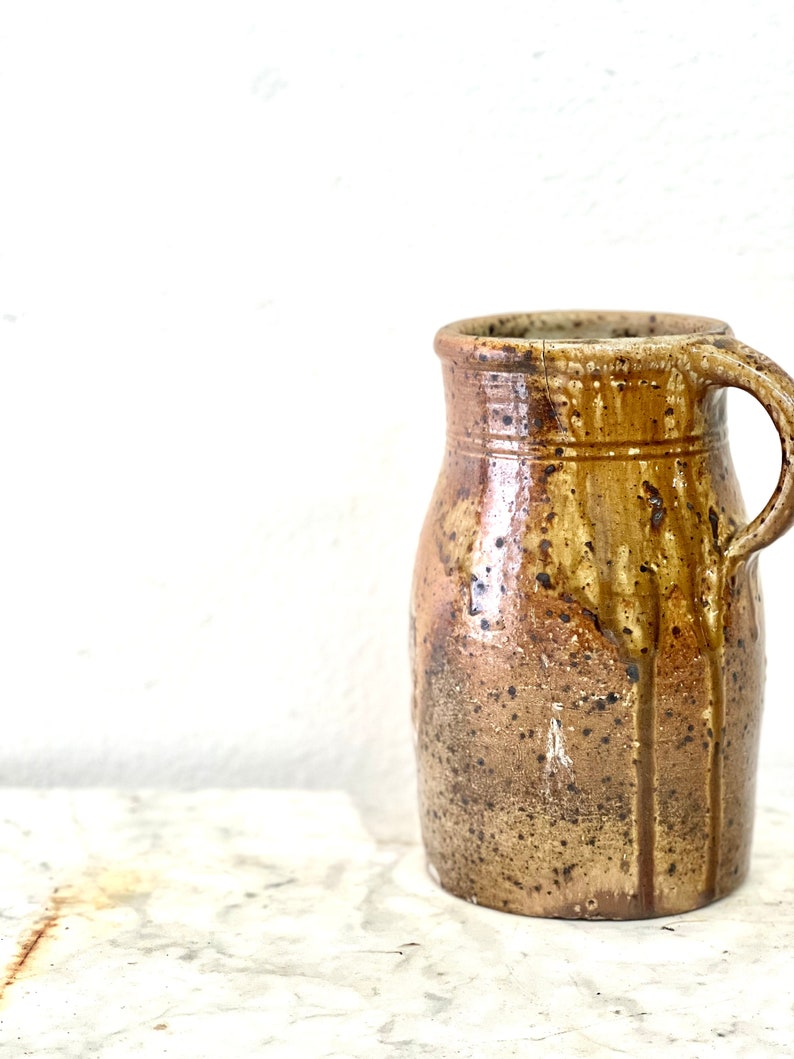 May include: A brown ceramic pitcher with a speckled glaze and a handle. The pitcher has a rounded body and a slightly flared lip.