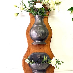 May include: A wall-mounted wooden plaque featuring two attached pewter vessels. The top vessel is a tall, rounded vase with a lion head relief, and the bottom is a wide, bowl-shaped basin with a lion head detail, both holding white flowers.