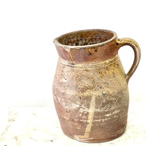 May include: A brown ceramic pitcher with a wide mouth and a handle. The pitcher has a rustic, handmade look and is glazed in a light brown color.