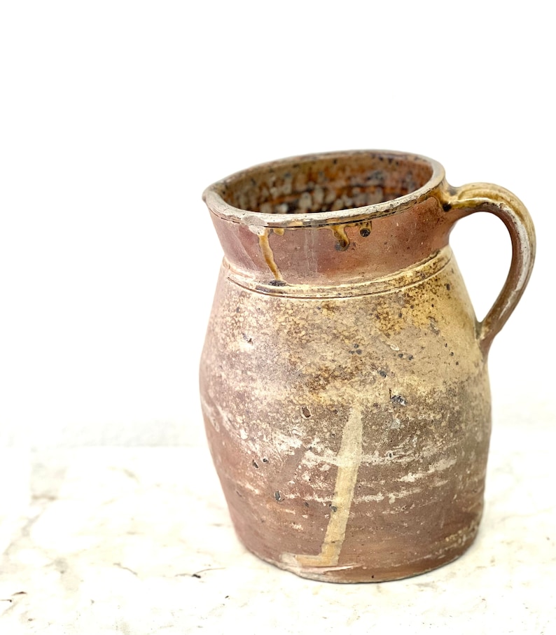 May include: A brown ceramic pitcher with a wide mouth and a handle. The pitcher has a rustic, handmade look and is glazed in a light brown color.