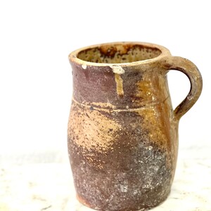 May include: A brown earthenware pitcher with a handle. The pitcher has a rustic, aged look with a mottled brown and tan glaze.