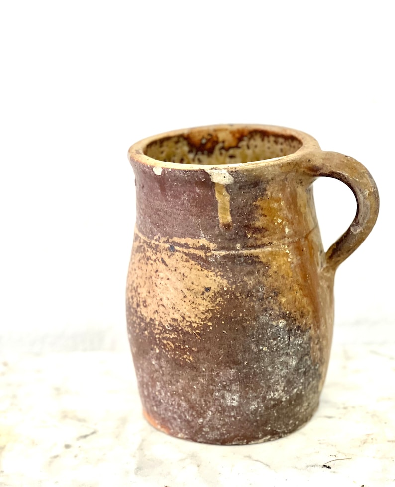 May include: A brown earthenware pitcher with a handle. The pitcher has a rustic, aged look with a mottled brown and tan glaze.