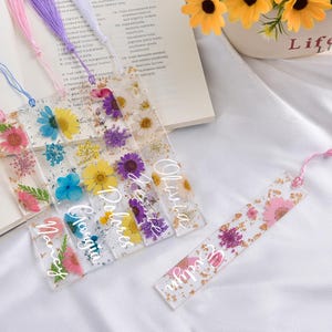 May include: Clear resin bookmarks with dried flowers and gold leaf. Each bookmark has a tassel and a name in white script. The flowers include pink, yellow, blue, and purple blooms.