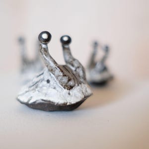 May include: Ceramic snail figurines with a textured, white and grey body and dark brown base. The snail's antennae have a dark brown tip. The figurines are on a white surface.