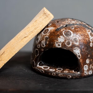 May include: A dome-shaped ceramic incense holder with a brown and white speckled glaze featuring a large arched opening, positioned next to a rectangular piece of light tan wood used for smudging or burning as incense.