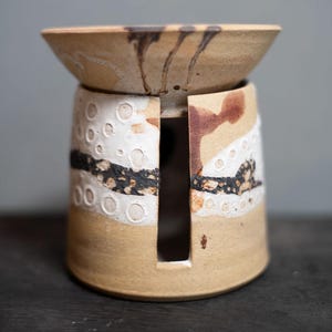 May include: A cylindrical ceramic oil burner with a wide, flared top bowl. The exterior features a tan base with a white textured band containing circular indentations, a horizontal black speckled stripe, and brown drip accents on the upper rim.