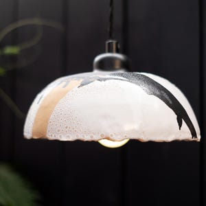 May include: A dome-shaped ceramic pendant light fitting featuring a white glazed surface with a textured, bubbly finish. The shade is decorated with abstract black and tan brushstroke patterns and hangs from a black cord and fixture mount.