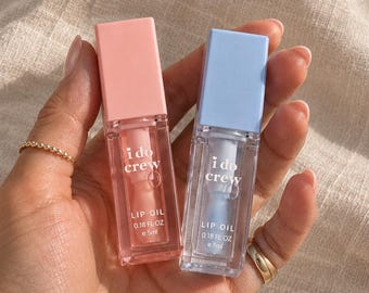 May include: Two rectangular lip oil tubes with clear bodies and coloured caps, one pastel pink and one pastel blue. Each tube is labelled with the text i do crew LIP OIL 0.18 FL OZ e 5ml in white lettering on the front surface.