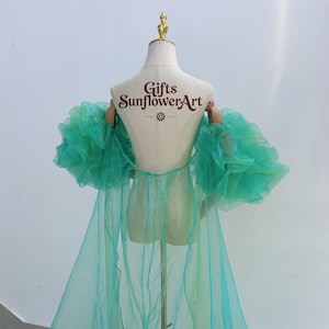 May include: A sheer, turquoise-green robe with voluminous ruffled sleeves is displayed on a mannequin. The back of the mannequin has the text "Gifts SunflowerArt". The robe's fabric drapes down, creating a flowing effect. The overall design is elegant and airy.