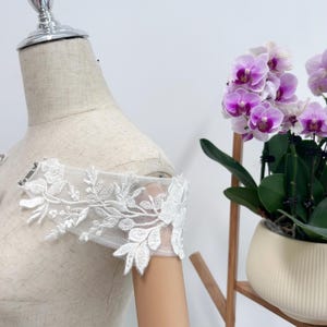 Könnte beinhalten: Weißes florales Spitzenärmeldetail. Die zarte Spitze weist komplizierte Blumen- und Blättermuster auf. Der Ärmel ist an einer Schaufensterpuppe befestigt, die das Design zeigt. Eine Topf-Orchidee mit lila Blüten befindet sich im Hintergrund.