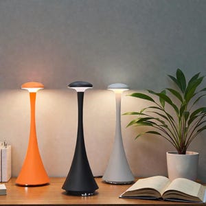 Puede incluir: Tres lámparas de mesa modernas en naranja, negro y blanco. Cada lámpara tiene una base cónica y un tallo delgado que conduce a una luz plana y circular. Las lámparas están sobre una superficie de madera, con una planta y un libro abierto en el fondo.