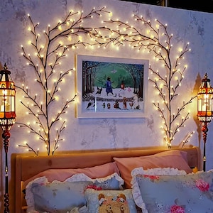 May include: A bedroom scene featuring a wooden bed frame, adorned with pink and blue floral pillows. Decorative branches with warm white LED lights are arranged on the wall, framing a framed picture. Two ornate floor lamps with amber glass panels flank the bed.