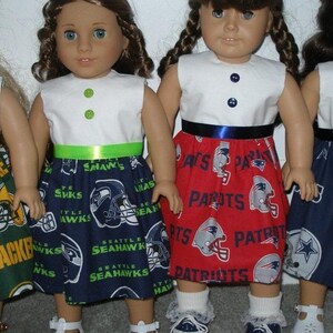 May include: Dolls wearing handmade outfits. One doll is wearing a white top with a green ribbon and a blue skirt with Seattle Seahawks logos. Another doll is wearing a white top with a blue ribbon and a red skirt with New England Patriots logos.