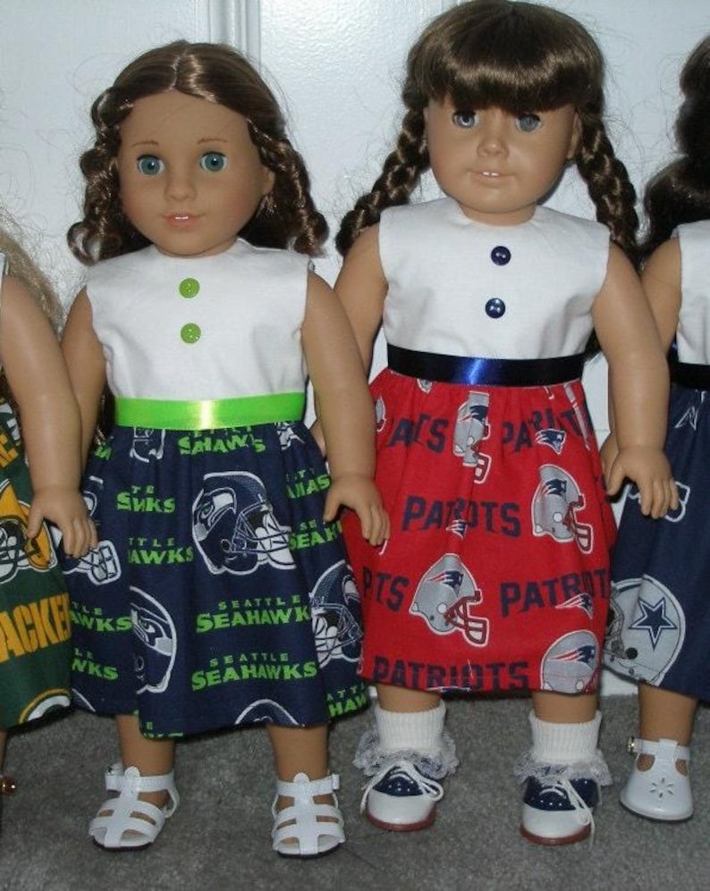 May include: Dolls wearing handmade outfits. One doll is wearing a white top with a green ribbon and a blue skirt with Seattle Seahawks logos. Another doll is wearing a white top with a blue ribbon and a red skirt with New England Patriots logos.