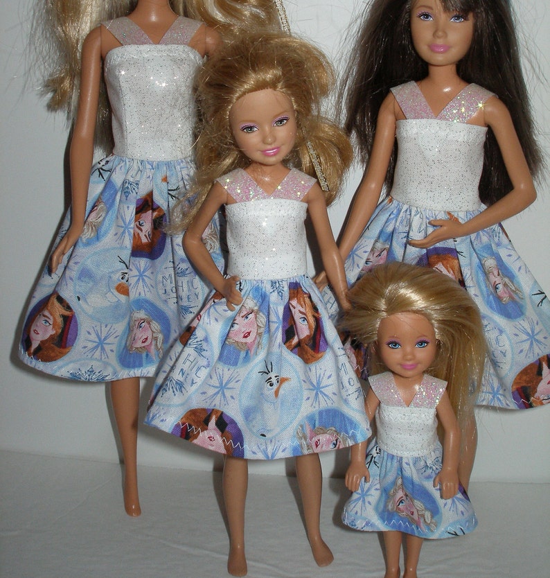 Puede incluir: Tres mu&ntilde;ecas con vestidos blancos con purpurina y faldas azules con un estampado de Frozen. Las mu&ntilde;ecas est&aacute;n de pie sobre una superficie blanca.