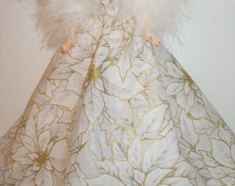 May include: A doll wearing a white gown with a gold floral pattern. The dress has a full skirt and is adorned with a white feathered boa. The doll's arms are visible, and it has red nail polish.