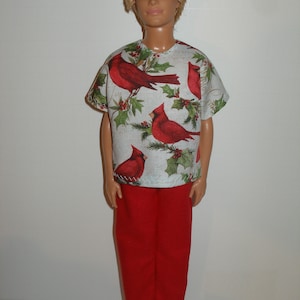 May include: A doll wearing a short-sleeved shirt and red pants. The shirt is white with a pattern of red cardinals and green holly leaves. The red pants are a solid color.
