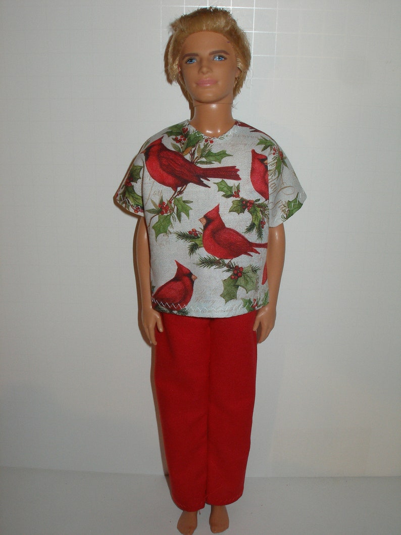 May include: A doll wearing a short-sleeved shirt and red pants. The shirt is white with a pattern of red cardinals and green holly leaves. The red pants are a solid color.