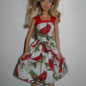 May include: A doll wearing a red and white dress with a cardinal bird print. The dress has a red bodice and a flared skirt with a pattern of red cardinals, holly, and the text "Holiday Greetings".
