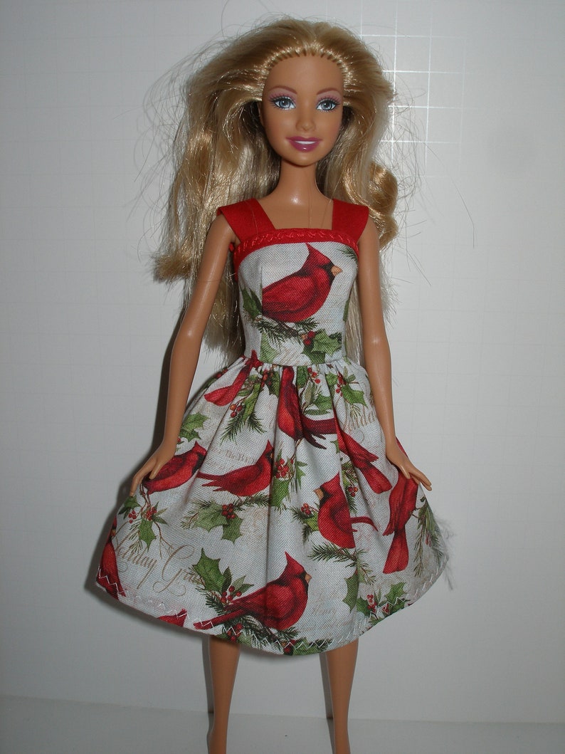 May include: A doll wearing a red and white dress with a cardinal bird print. The dress has a red bodice and a flared skirt with a pattern of red cardinals, holly, and the text "Holiday Greetings".