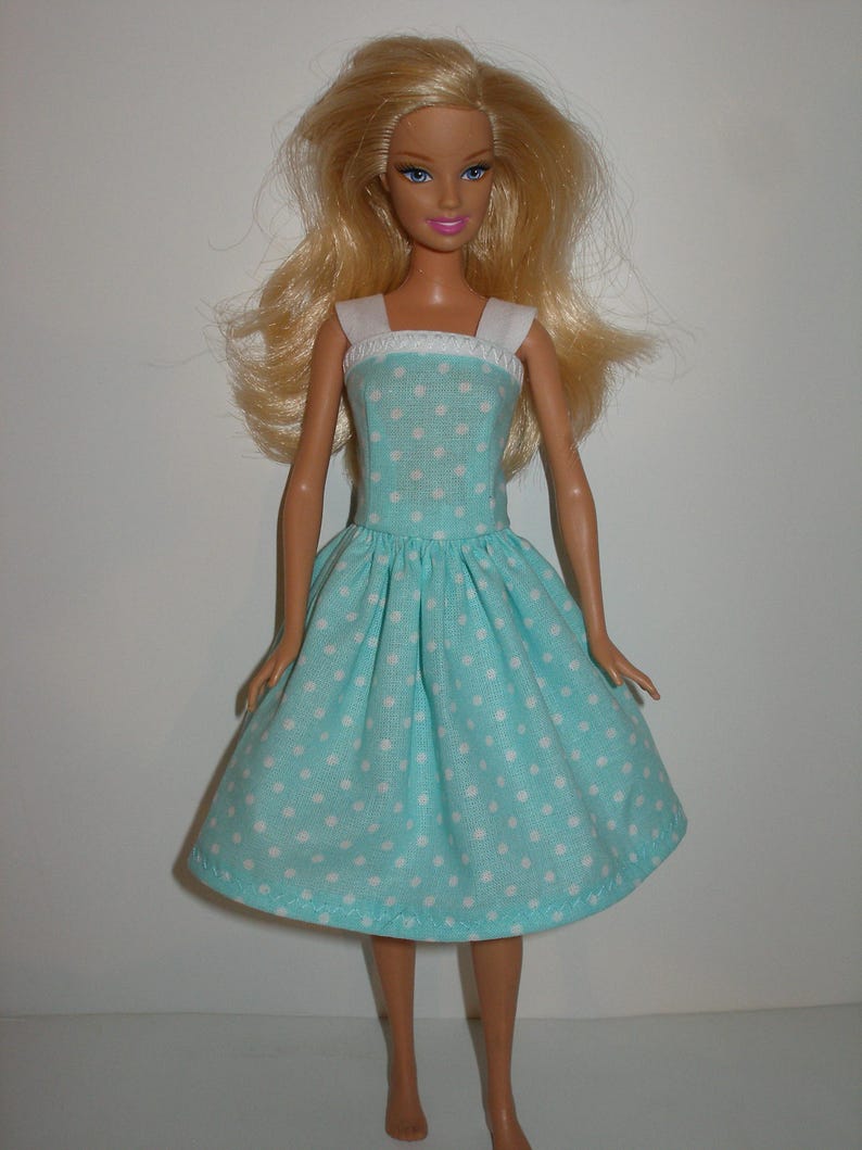 K&ouml;nnte beinhalten: Eine Puppe mit blonden Haaren und blauen Augen, die ein hellblaues Kleid mit wei&szlig;en Punkten tr&auml;gt. Das Kleid hat einen wei&szlig;en Ausschnitt und Tr&auml;ger. Die Puppe steht vor einem schlichten wei&szlig;en Hintergrund.