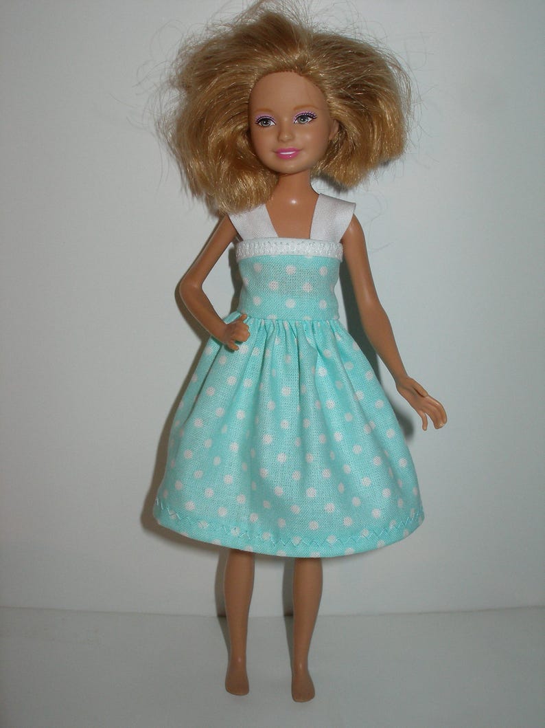 K&ouml;nnte beinhalten: Puppe in einem hellblauen Kleid mit wei&szlig;en Punkten. Das Kleid hat ein wei&szlig;es Oberteil und einen ausgestellten Rock. Die Puppe hat blondes Haar und steht vor einem wei&szlig;en Hintergrund.