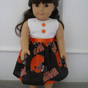May include: A doll wearing a white sleeveless top with two orange buttons and a black skirt. The skirt features orange football helmets and the words "CLEVELAND BROWNS". An orange ribbon accents the waist. The doll is wearing white shoes.
