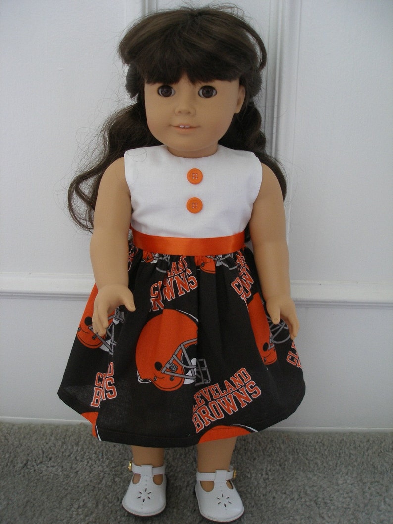 May include: A doll wearing a white sleeveless top with two orange buttons and a black skirt. The skirt features orange football helmets and the words "CLEVELAND BROWNS". An orange ribbon accents the waist. The doll is wearing white shoes.