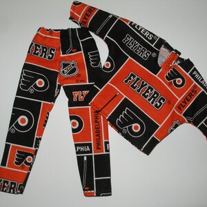 May include: A black and orange pajama set with the Philadelphia Flyers logo and team name. The set includes a long-sleeved shirt and pants.