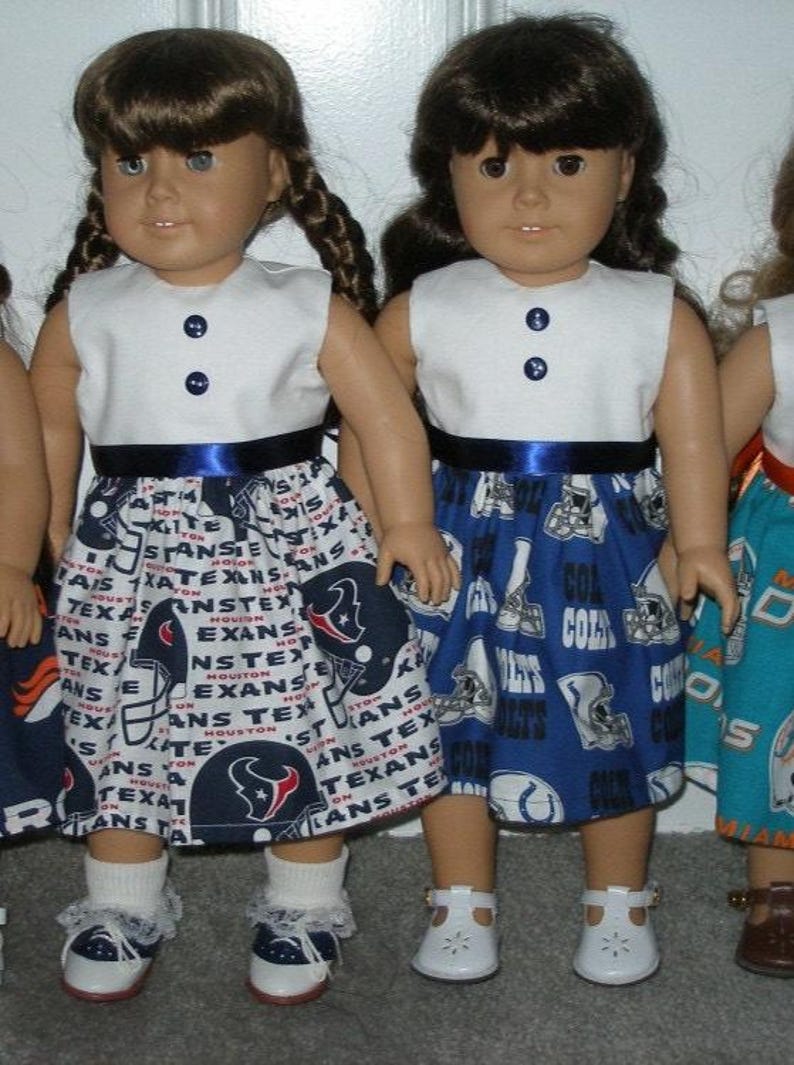May include: Two dolls wearing white sleeveless tops with navy blue buttons and a navy blue ribbon belt. One doll has a skirt with a Houston Texans football theme, and the other has a skirt with a Colts theme. Both dolls wear white shoes.
