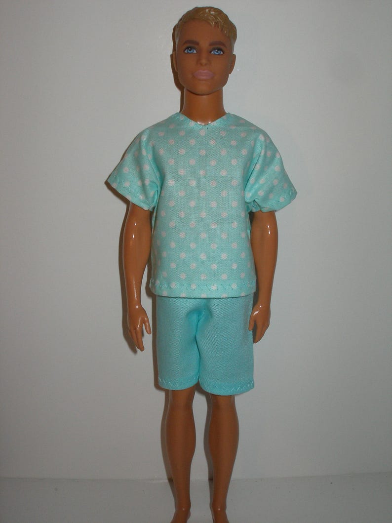 K&ouml;nnte beinhalten: Eine Puppe in einem hellblauen Pyjama-Set. Das kurz&auml;rmelige Oberteil ist mit wei&szlig;en Punkten gemustert. Die passenden Shorts vervollst&auml;ndigen das Outfit. Die Puppe hat blondes Haar und blaue Augen.