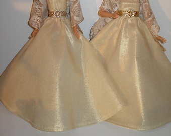 May include: Two dolls wearing matching gold and white gowns with lace shawls. The gowns have a shimmery finish and are strapless. Each doll has a gold belt with a jeweled buckle.