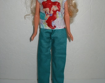 May include: A doll wearing a white sleeveless shirt with a printed image of Ariel from The Little Mermaid. The doll is also wearing teal pants.